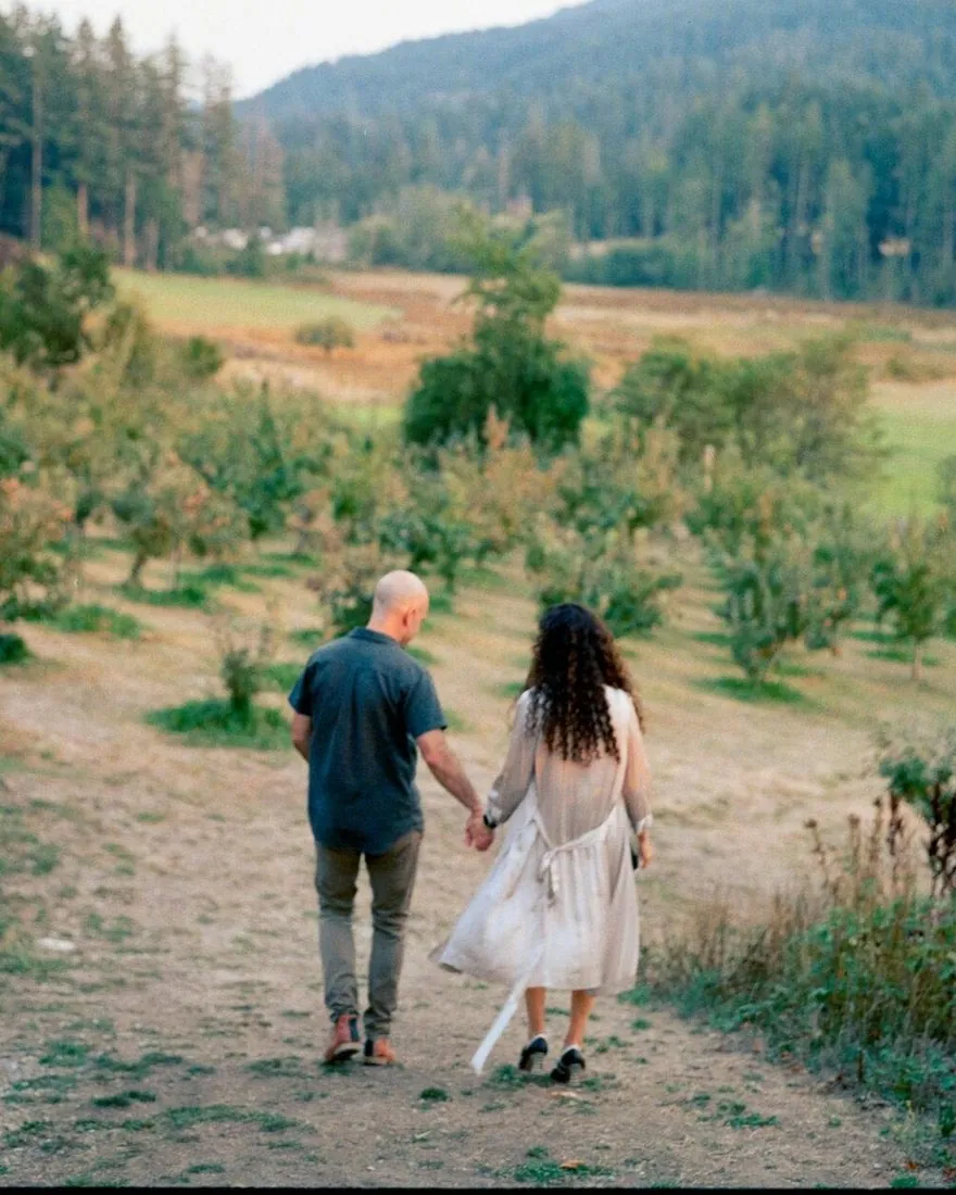 Ethan Wilding and his wife Bahareh walking hand in hand along a path through a Salt Spring orchard, forested ridge behind them