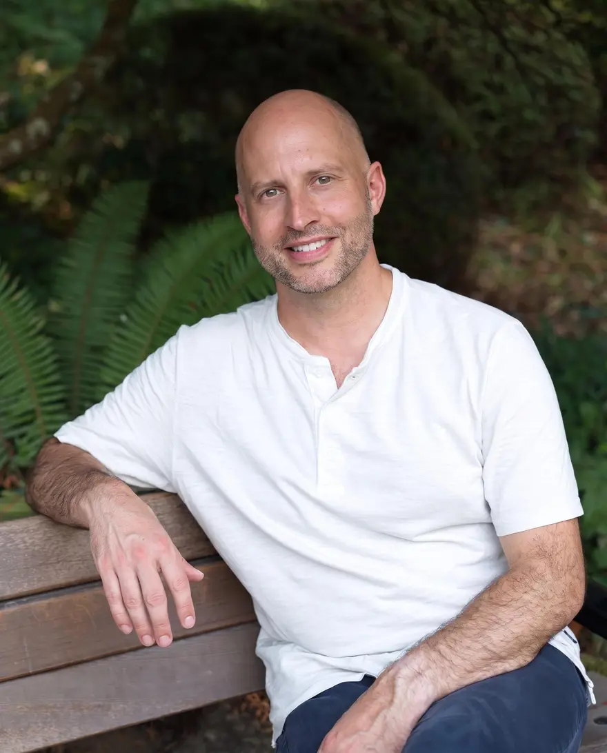 Ethan Wilding sitting on a wooden bench in a Salt Spring garden, sword ferns and green shade behind him, smiling toward the camera